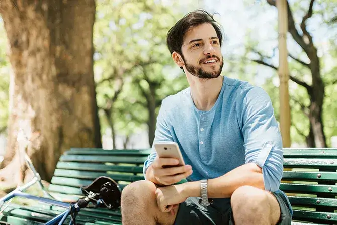 A man smiles and holds his phone while sitting on a park bench