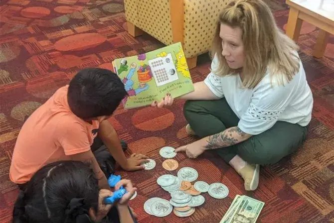 Financial educator Tori Filas sits on the floor reading a book to two children with large play money on the floor between them.