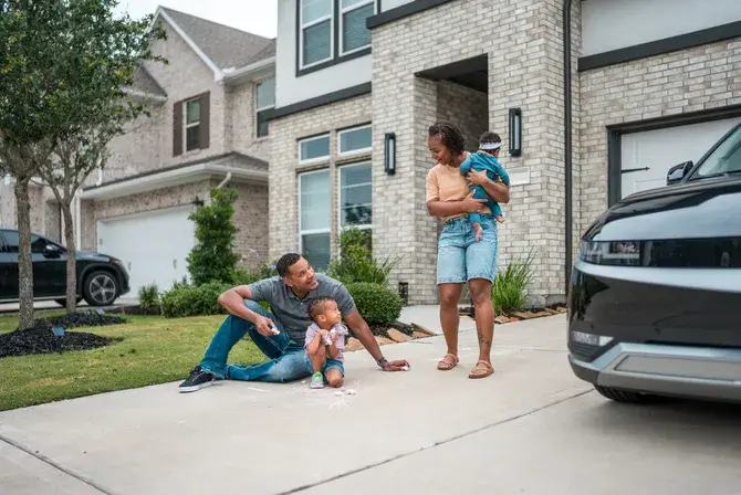 A smiling family engages in outdoor fun by their house, with a mother holding her child, as the father draw with chalk with their toddler near their parked car in the driveway.