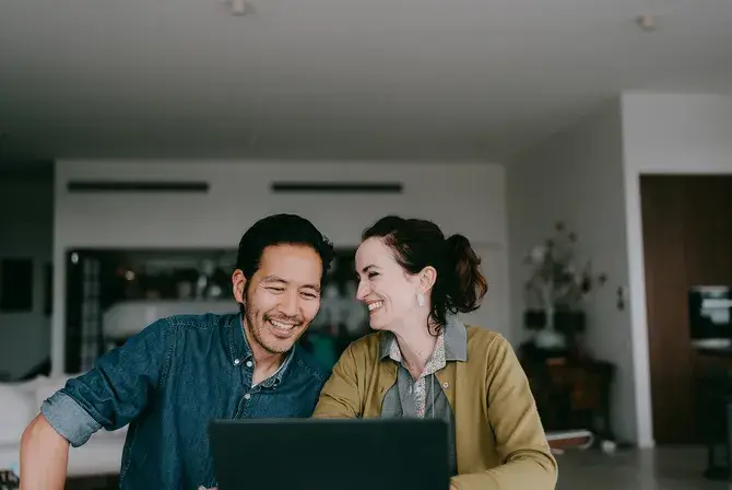 A man and woman smiling together while looking at a laptop screen in their kitchen.