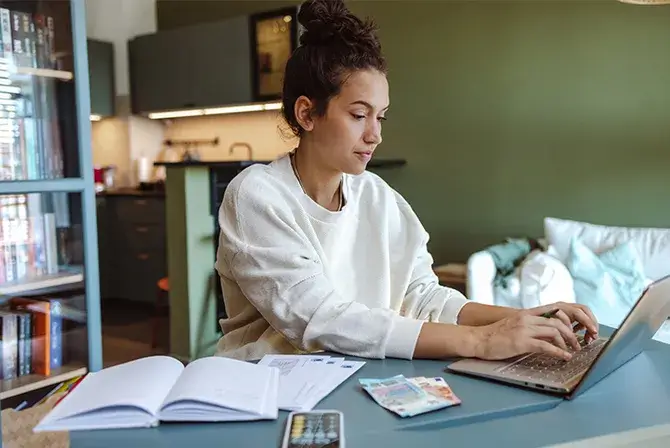 Young woman seated at a table working on her laptop. Money, books and a calculator are visible in the foreground and indicate she could be working on a budget.