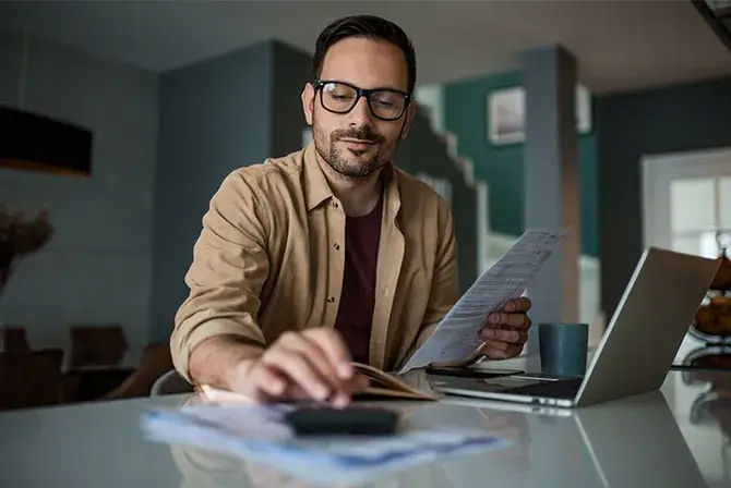 A man sits at a desk with an open lap and smiles confidently as he types into a calculator, holding a piece of paper in his other hand.