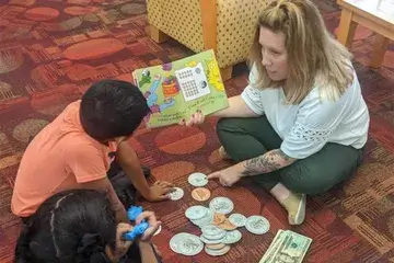 Financial educator Tori Filas sits on the floor reading a book to two children with large play money on the floor between them.
