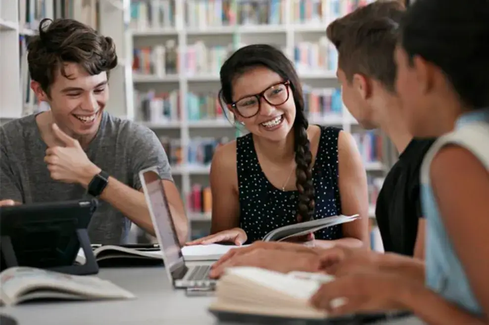 A group of teenagers smiles sitting in a library, surrounded by book and computers on the table.