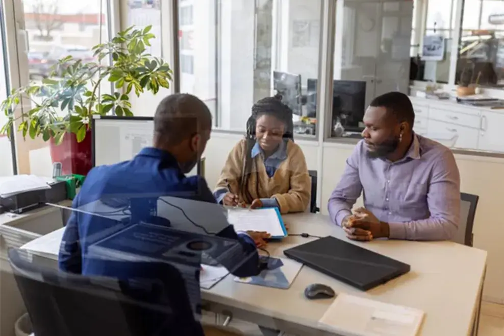 A couple sits at a desk reviewing paperwork with a car dealership employee