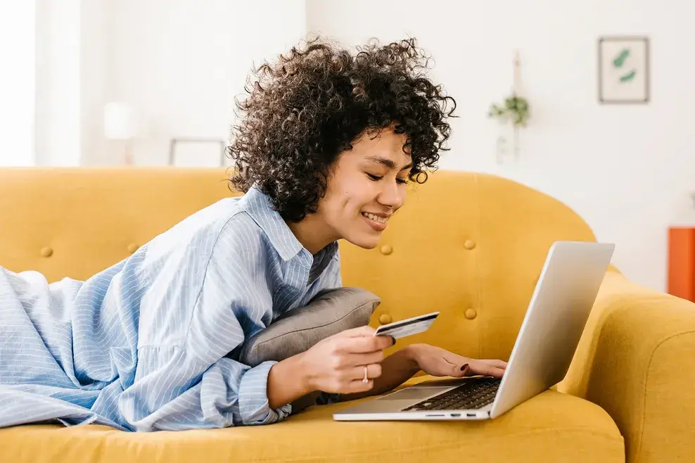 Smiling woman holding credit card using laptop lying on sofa in living room at home