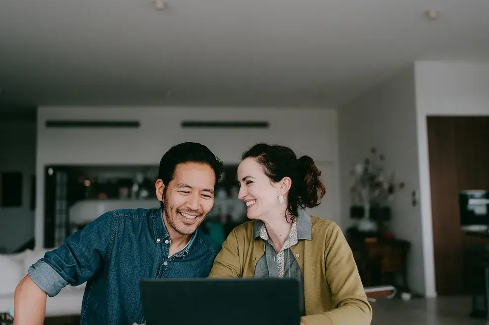 A man and woman smiling together while looking at a laptop screen in their kitchen.