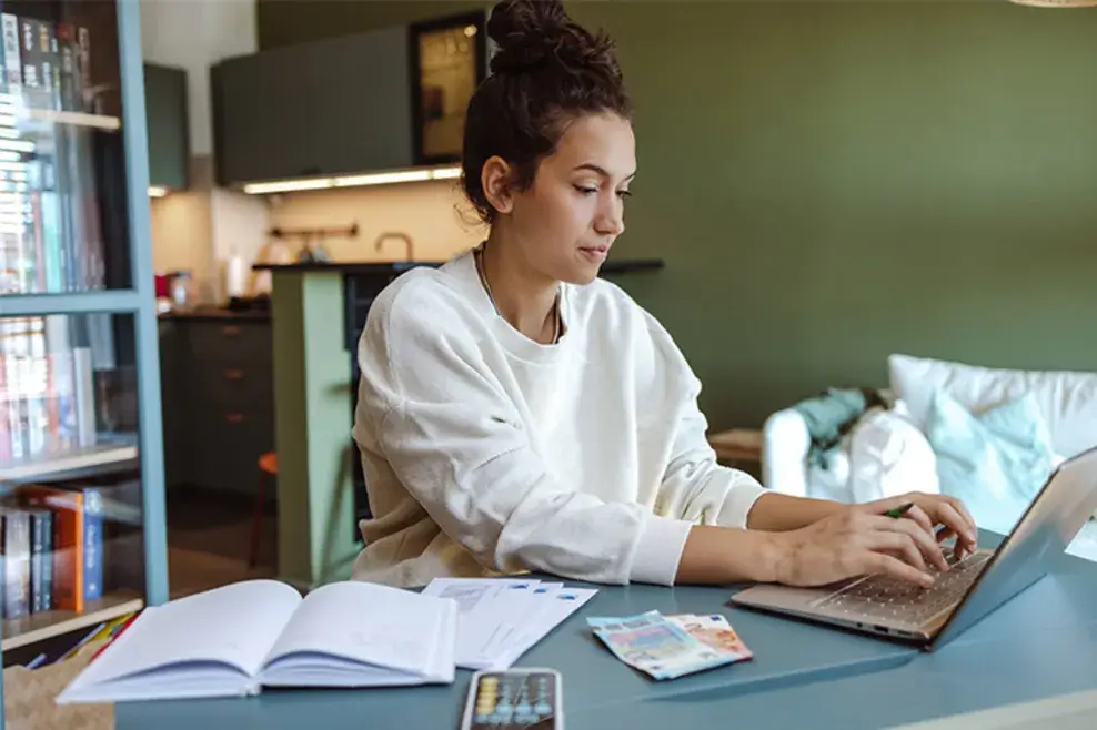 Young woman seated at a table working on her laptop. Money, books and a calculator are visible in the foreground and indicate she could be working on a budget.