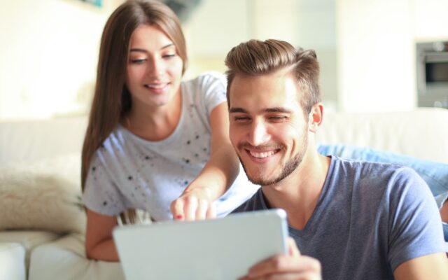 Smiling young couple sit together in living room woman points to tablet in mans hands 978b8d064138018a1dfad71b9882dd21