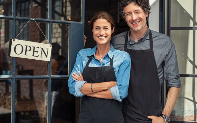 Business owning couple wearing shop aprons stand outside door with 22 Open22 sign 978b8d064138018a1dfad71b9882dd21