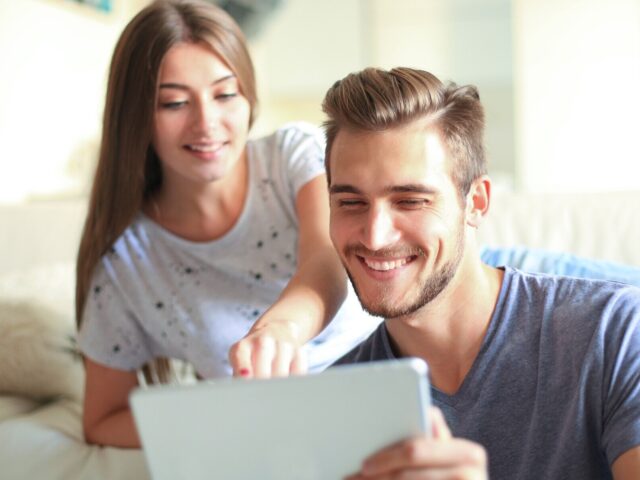 Smiling young couple sit together in living room woman points to tablet in mans hands 978b8d064138018a1dfad71b9882dd21