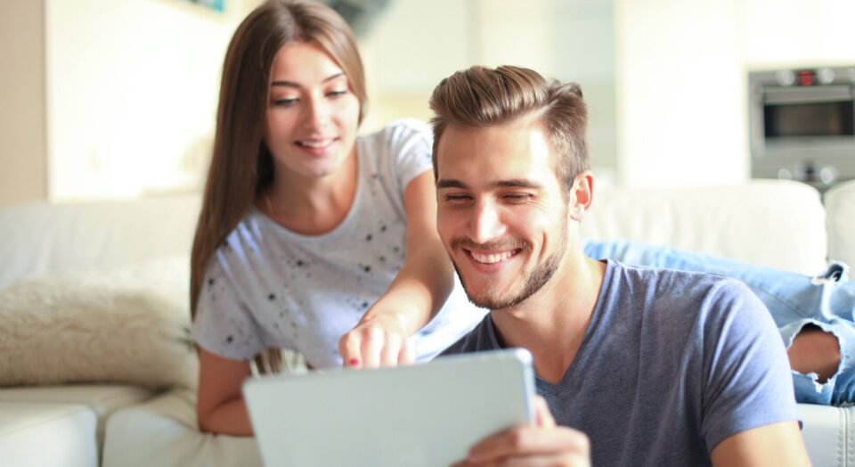 Smiling young couple sit together in living room woman points to tablet in mans hands 978b8d064138018a1dfad71b9882dd21