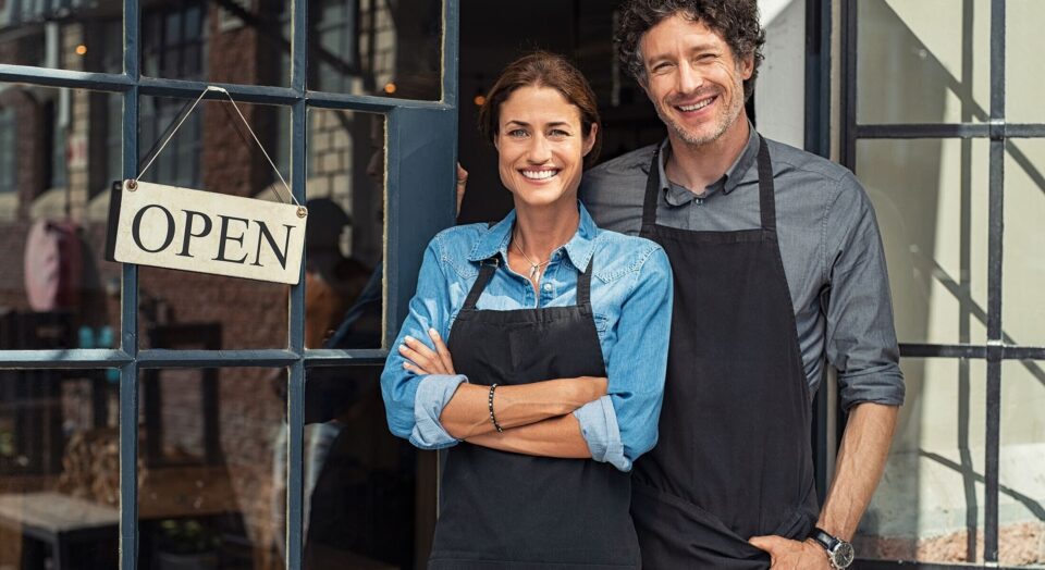 Business owning couple wearing shop aprons stand outside door with 22 Open22 sign 978b8d064138018a1dfad71b9882dd21