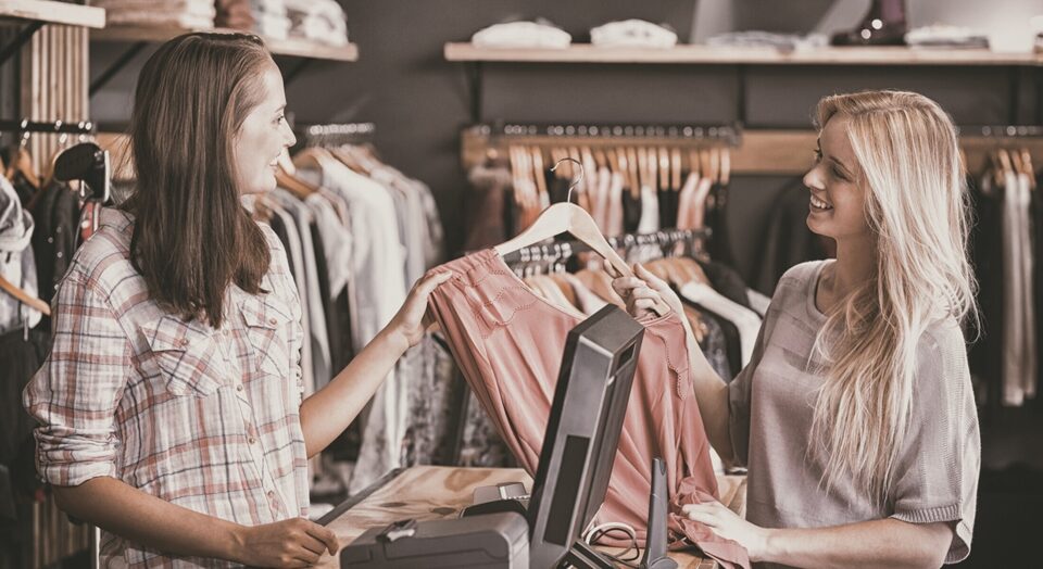 Young woman at clothing store checkout hands shirt to sales associate 978b8d064138018a1dfad71b9882dd21